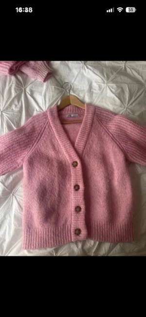 Rosa stickad kofta med knappar - Supermysig rosa stickad kofta från Zara med v-ringad hals och fyra bruna knappar framtill. Koftan har långärmade raglanärmar och breda ribbade muddar vid ärmslut och nederkant. Perfekt för att addera lite färg till din garderob. Lite nopprig