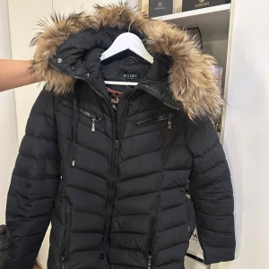 Svart pufferjacka med päls från Hollies - Snygg svart pufferjacka från Hollies med fluffig päls på huvan och markerat bälte i midjan. Jackan har flera dragkedjor framtill, quiltad design och långa ärmar. Perfekt för kalla dagar med sin varma och stilrena look.