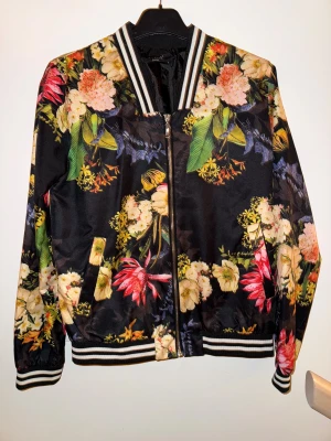 Blommig bomberjacka med dragkedja - Säljer en svart bomberjacka från NA-KD med färgglatt blommigt mönster och vita ränder på krage, ärmslut och nederkant. Jackan har dragkedja framtill och två fickor. Perfekt för dig som vill sticka ut med en cool och unik stil.