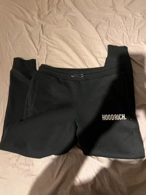 Svarta mjukisbyxor från Hoodrich - Svarta mjukisbyxor från Hoodrich med vit logga tryckt på vänster ben. Byxorna har snörning i midjan, ribbade muddar vid bensluten och praktiska sidofickor. Materialet är mjukt och bekvämt, perfekt för chill eller streetwear. Snygg och enkel design. Längd från midja till fot 100cm
