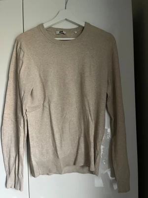 Beige stickad tröja från WE, S - En stilren beige stickad tröja från WE i storlek S. Tröjan har rund hals och ribbade muddar vid ärmslut. Perfekt för dig som gillar enkel och clean look med mjukt material.
