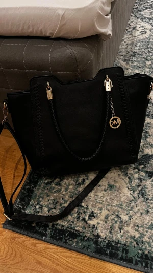 Svart Michael Kors axelväska i skinn - Snygg svart axelväska från Michael Kors med flätade handtag och guldfärgade detaljer. Väskan har MK-logga i metall, dragkedja upptill och flera fack inuti. Materialet är skinn och designen är stilren med dekorativa sömmar längs kanterna.