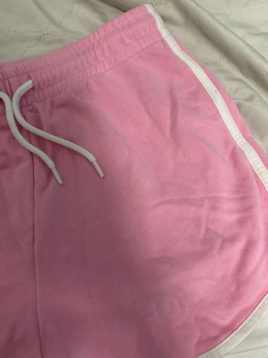 Rosa sportiga shorts med vita detaljer - Säljer ett par ljusrosa shorts från Cubus i storlek 158/164. De har vita snören i midjan och vita kantband längs sidorna. Modellen är loose och har elastisk midja med snörning, perfekt för chill eller träning. Materialet är mjuk bomull som känns skönt mot huden.