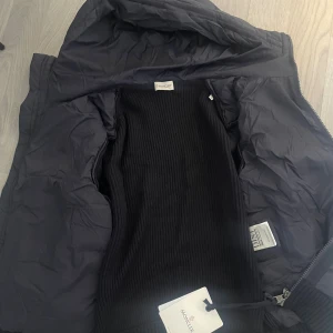  Moncler cardigan marin blå - Säljer en svart pufferjacka från Moncler med stickade ärmar och quiltad kropp. Jackan har huva, dragkedja framtill och en liten ficka med dragkedja på ena ärmen. Perfekt för kyliga dagar och riktigt snygg streetstil.  Helt ny fick den 31 okt 2025
