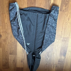 Svart vindjacka från Adidas - Svart vindjacka från Adidas med diskret mönstrade ärmar och reflekterande ränder längs sidorna. Jackan har dragkedja framtill, huva och Adidas-logga på bröstet. Tillverkad i lätt polyester, perfekt för blåsiga dagar. Är storlek 152 