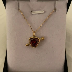 Guldigt halsband med röd hjärthänge - Superfint halsband i guldton med ett hjärtformat rött hänge och pil-detalj. Kedjan är tunn och elegant, och hänget har en gnistrande röd sten i mitten. Perfekt för dig som gillar romantiska smycken med lite extra attityd. Rostfri 