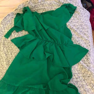 Grön omlottklänning med volanger - Superfin grön klänning med omlottkänsla och volangdetaljer. Klänningen har en axelbandslös design med knyt på ena axeln och markerad midja. Perfekt för dig som vill sticka ut med en färgstark look. Materialet känns lätt och passar bra till varmare dagar.