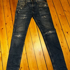 Replay Sustainable Lab 28/32 - Säljer ett par Replay jeans i blå tvätt med slitna detaljer och skinny passform. Klassisk femficksmodell med låg midja och smala ben. Jeansen är tillverkade i stretchig denim för extra komfort och har snygga slitningar framtill och på bakfickorna.