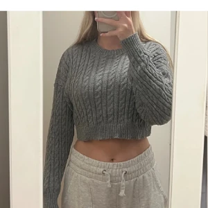 Grå croppad stickad tröja - Säljer en grå croppad stickad tröja med kabelstickat mönster. Tröjan har rund halsringning och långa ärmar. Perfekt för dig som gillar en trendig och avslappnad stil.