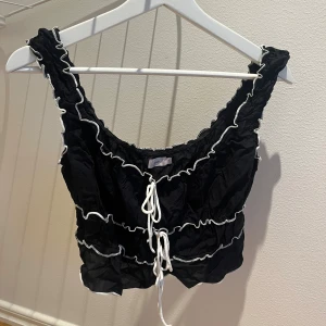 Svart croppad topp med volang och snörning - Svart croppad linnetopp med vita volangdetaljer och snörning framtill. Toppen har breda axelband och är ärmlös, med en lätt rynkad struktur och kontrasterande vita sömmar som ger en cool och trendig look.