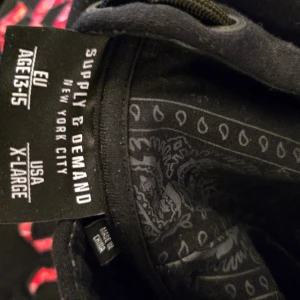 Svart hoodie Supply & Demand X-Large - Svart hoodie från Supply & Demand med stor röd logga i gotisk stil på bröstet och vita detaljer. Hoodien har snörning i huvan och coola paisleymönstrade detaljer på insidan och vid fickorna. Mjuk och skön bomullsmix, perfekt för streetwear.