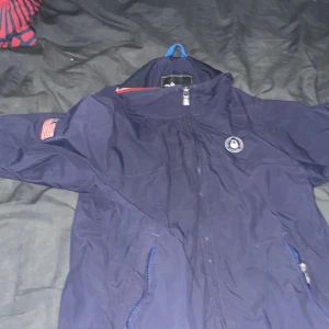 Marinblå vindjacka Sail Racing 160 - Snygg marinblå vindjacka från Sail Racing med dragkedja, broderad USA-flagga på ärmen och trycket 'USA 77' på ryggen. Jackan har sportig känsla, meshfoder och detaljer i rött och vitt vid muddarna. Perfekt för dig som gillar stilren och sportig look.