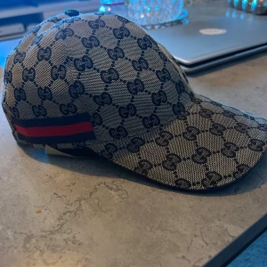 Gucci keps med GG-mönster och röd-blå detalj - Snygg keps från Gucci med klassiskt GG-mönster i beige och svart. Kepsen har en böjd skärm och en röd-blå rand på sidan som ger extra lyxig vibe. Tillverkad i bomull med justerbar rem baktill för perfekt passform. Perfekt för dig som vill sticka ut med designerstil.