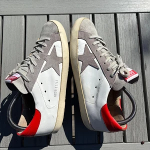 Golden Goose Super-Star sneakers – klassiker! - Storlek: 41  Säljer mina Golden Goose Super-Star i vitt skinn med grå mockadetaljer och röd häl. Den ikoniska modellen med stjärnan på sidan ⭐️  ✔️ Nypris ca 5 000 kr ✔️ Fräscha sulor, mycket mönster kvar ✔️ Golden Goose görs med “vintage look” från start – så de ska se lite slitna ut!  Snygga till allt från jeans till kostym byxor .