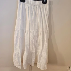 Vit långkjol från Lindex - Luftig och snygg vit långkjol från Lindex med bred resår i midjan och flera volangpartier. Kjolen är gjord i mjuk bomull och har en avslappnad passform som ger en skön och somrig vibe.