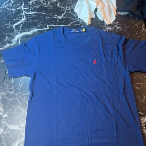 Blå t-shirt från Polo Ralph Lauren - Snygg marinblå t-shirt från Polo Ralph Lauren med klassisk röd broderad logga på bröstet. Modellen är kortärmad och har rund hals. Materialet är mjuk bomull som känns skönt mot huden. Perfekt för dig som gillar stilrena och ikoniska plagg.