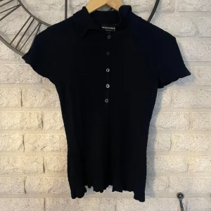 Svart stickad topp från Emporio Armani - Svart stickad topp från Emporio Armani med korta ärmar, krage och knappar framtill. Ribbad struktur och lätt figurnära passform ger en clean och stilren look. Perfekt för dig som gillar klassiska designer med en modern twist.
