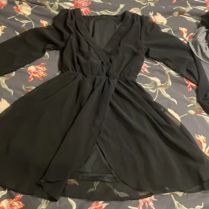 Svart omlottklänning med v-ringning - Snygg svart kortklänning perfekt för nyår och  med omlott-design och v-ringad hals. Klänningen har långa ärmar och ett lätt, flödande tyg som ger en elegant siluett. Perfekt för dig som gillar stilrena och trendiga plagg med lite extra attityd.