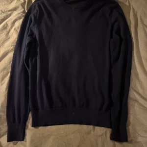 Mörkblå stickad tröja i ull - En stilren mörkblå stickad tröja med rund hals och långa ärmar. Tröjan har ribbade muddar vid ärmslut och nederkant, vilket ger en klassisk och clean look. Perfekt för lager-på-lager under kyliga dagar, ifrån uniqlo samt gjord i merino ull.