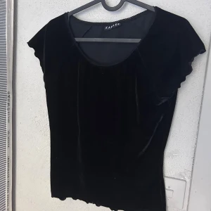 Svart sammetsblus från Kaffe - Svart sammetsblus från Kaffe med rund halsringning och korta, lätt volangiga ärmar. Toppens material ger en lyxig känsla och den har en mjuk, följsam passform. Perfekt för dig som gillar en stilren men ändå elegant look.