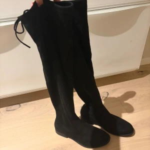 Svarta overknee boots i mocka Tiamo - Säljer ett par svarta overknee boots från Tiamo. Aldrig använda endast testade inne. med platt sula och rund tå. Bootsens ovandel är i mjuk mocka och de har snörning upptill för att sitta snyggt på benet. Perfekt för dig som vill ha en stilren och trendig look under höst och vinter.