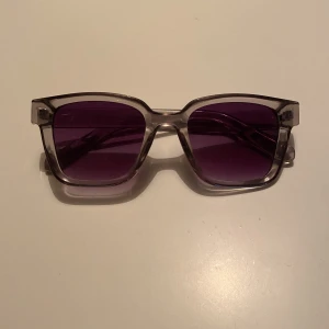 Transparenta solglasögon med lila glas - Snygga solglasögon med transparent, fyrkantig båge och lila tonade glas. Ramen är i plast och ger en modern vibe, perfekt för att sticka ut i mängden. Passar dig som gillar en stilren men ändå färgstark accessoar.