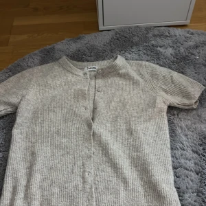 Beige stickad kofta från J'aime - Supermysig beige stickad kofta från J'aime med korta ärmar och ribbad struktur. Koftan har rund halsringning och knäppning framtill med matchande knappar. Perfekt att slänga på sig över en t-shirt eller linne.