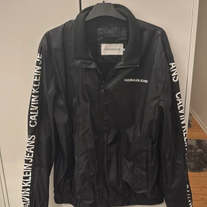 Svart vindjacka från Calvin Klein Jeans - Svart vindjacka från Calvin Klein Jeans med vit logga på bröstet och längs båda ärmarna. Jackan har dragkedja framtill, elastiska muddar och en sportig look. Tillverkad i lätt polyester som passar perfekt för blåsiga dagar.