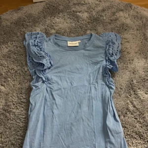 Blå blus med volangärmar Fabienne Chapot - Supersöt ljusblå blus från Fabienne Chapot i bomull med volangiga, spetsprydda kortärmar. Perfekt för dig som vill ha något unikt och luftigt till sommaren. Snygg rund halsringning och romantiska detaljer på ärmarna.