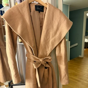 Beige kappa från Xoul Edition med bälte - Snygg beige kappa från Xoul Edition med bred krage och knytbälte i midjan. Jackan har lång ärm, slät yta och en klassisk, stilren look. Perfekt för dig som vill ha en trendig och tidlös ytterjacka med extra schysst passform.