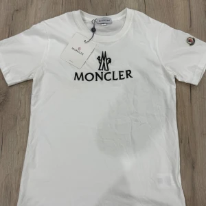 Vit Moncler t-shirt med logga - Vit t-shirt från Moncler i bomull med svart broderad logga på bröstet och klassisk patch på ärmen. T-shirten har rund halsringning och korta ärmar, perfekt för en clean och stilren look.