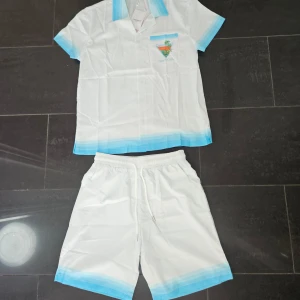 Casablanca Tennis Club vita shorts - Snygga vita shorts från Casablanca med elastisk midja och snörning. De har en fräsch blå kant längst ner och är gjorda i ett lätt material som passar perfekt för tennis eller sommarhäng. Matchar skjortan med samma blå detaljer och tryck.