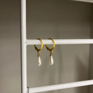 Guldörhängen med droppformad pärla - Snygga örhängen i guldton med droppformade vita pärlor som hänger från små ringar. Perfekt för dig som gillar klassisk och elegant stil med en modern twist. Passar till alla tillfällen och ger en lyxig känsla till din look. I rostfritt stål och handgjort