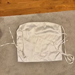 Vit satin bandeau tubtopp med snörning - Säljer en vit bandeau tubtopp i satin med snörning i sidorna och helt öppen rygg. Toppen är axelbandslös och har en slät, glansig yta som ger en lyxig känsla. Perfekt för dig som vill ha en enkel men snygg topp till din outfit.