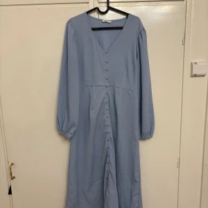 Ljusblå långklänning från Happy Holly - Säljer en ljusblå långklänning från Happy Holly med v-ringning och långa ärmar. Klänningen har knappar längs hela framsidan och ett lätt strukturerat tyg i polyester. Perfekt för dig som gillar stilrena och feminina detaljer.