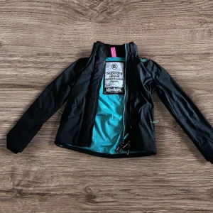 Svart vindjacka Superdry Japan M - Storlek M men sitter som S - Svart vindjacka från Superdry Japan med turkos meshfoder och coola detaljer som ribbad krage och dragkedjor. Jackan har logotyp på bröstet och orange Superdry-märke vid ärmen. Perfekt för dig som gillar streetwear och snygg funktion.