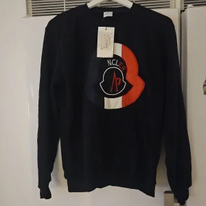 Svart sweatshirt - Svart sweatshirt i storlek L med stort broderat Moncler-logo på bröstet i blått, vitt och rött. Tröjan har rund hals, långa ärmar och ribbade muddar. Perfekt för dig som gillar streetwear och vill ha en stilren look med exklusiv känsla. Passar även M. Helt ny!!