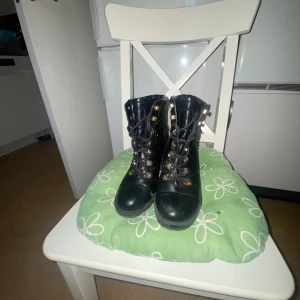 Svarta kängor med klack från Guess - Säljer ett par svarta kängor från Guess med hög klack och snörning. Skorna har guldfärgade detaljer. Rund tå och stabil platt sula gör dem både snygga och bekväma. Perfekt för dig som gillar edgy stil. Använda