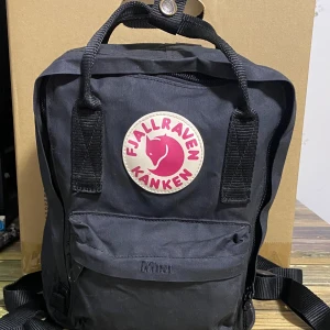 Svart Fjällräven Kånken ryggsäck - Klassisk svart Fjällräven Kånken ryggsäck med ikonisk rund logga framtill i rött och vitt. Ryggsäcken har ett stort huvudfack, framficka med dragkedja och två sidofickor. Tillverkad i slitstark syntet med justerbara axelremmar och bärhandtag upptill.