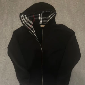 Svart Burberry vindjacka med huva - Svart vindjacka från Burberry med klassiskt rutigt foder i huvan och insidan. Jackan har dragkedja framtill, två fickor och en stilren look. Perfekt för dig som gillar ikoniska detaljer och snygg design.