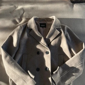 Beige kappa från ONLY med knappar - Snygg beige kappa från ONLY med dubbelknäppning och klassisk krage. Jackan har raka ärmar, två fickor framtill och stilrena svarta knappar. Perfekt för dig som gillar en clean och tidlös look.