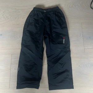 Didrikson Avan kids Navy täckbyxor från Didriksons - Svarta täckbyxor från Didriksons 1913 med broderad logga på benet. Byxorna har elastisk midja med snörning och praktisk röd hängare på insidan. Tillverkade i slitstark syntet, perfekta för kalla dagar utomhus.                        Aldrig anväd.