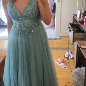 Ljusblå långklänning med pärlor - Säljer en ljusblå långklänning med v-ringad urringning och ärmlös design. Klänningen har ett glittrigt pärlbroderi upptill och ett flowigt, lager-på-lager tyg i kjolen. Perfekt för bal eller student! Materialet är troligtvis polyester med detaljer i spets och pärlor. Har även en slits vid ena benet. Inköpspris 4000