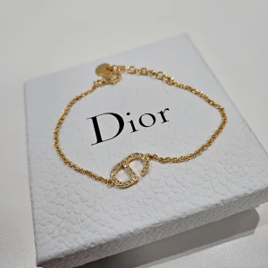 Guldarmband med Dior-logga - Snyggt och stilrent guldarmband från Dior med tunn kedja och ett glittrigt CD-emblem i mitten, dekorerat med små kristaller. Perfekt för dig som gillar exklusiva detaljer och vill ha något som sticker ut. Justerbar längd med klassisk låsning.