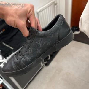Svarta Louis Vuitton sneakers i skinn - Svarta sneakers från Louis Vuitton med klassiskt monogrammönster över hela skon. Tillverkade i skinn med svarta snören och platt sula. Snygg och stilren modell som passar perfekt till streetwear eller en mer dressad look.