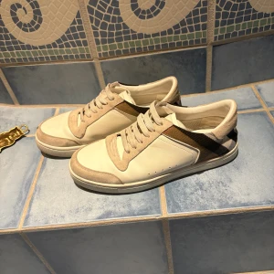 Burberry skor - Snygga vita sneakers från Burberry med klassisk rutig panel i beige, svart och brun på sidan och hälen. Skorna har snörning, rund tå och platt sula. Materialet är en mix av skinn och mocka, vilket ger en lyxig känsla. Perfekta för dig som gillar stilrena och exklusiva sneakers.