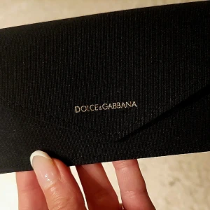 Svart glasögonfodral Dolce & Gabbana - Stilrent svart glasögonfodral från Dolce & Gabbana med guldfärgad logga på framsidan. Fodralet har en klassisk kuvertform och stängs med tryckknapp. Perfekt för att skydda dina glasögon med extra lyxig känsla.
