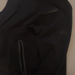 Svart vindjacka från Under Armour - Svart vindjacka från Under Armour i loose fit. Jackan har dragkedjor på fickorna och diskret logga på bröstet. Perfekt för dig som gillar sportig och clean stil. Materialet är lätt och känns smidigt att bära.