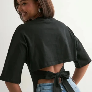 Svart croppad topp med knyt i ryggen - Svart croppad topp i bomull med halvlånga ärmar och öppen rygg som knyts med breda band. Enkel och stilren design med rund halsringning och loose passform. Perfekt för dig som vill ha en trendig och bekväm look.