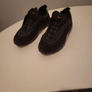Svarta Nike Air Max 97 med orange detaljer - Säljer ett par svarta Nike Air Max 97 sneakers med orange swoosh och detaljer. Skorna har en vågig design, synlig air-sula och snörning. Ovandelen är i syntet och textil, med en cool svart look och orange kontrast. Perfekta för dig som gillar streetstyle. Ser nya ut, lite småsvanker på gummit de svarta se sista bild, syns inte när skorn är på